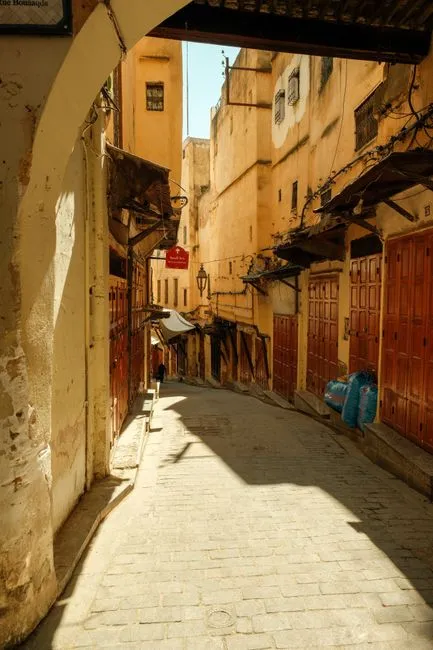 A narrow sunlit alley in Fes el-Bali medina with traditional wooden doors and stone walls